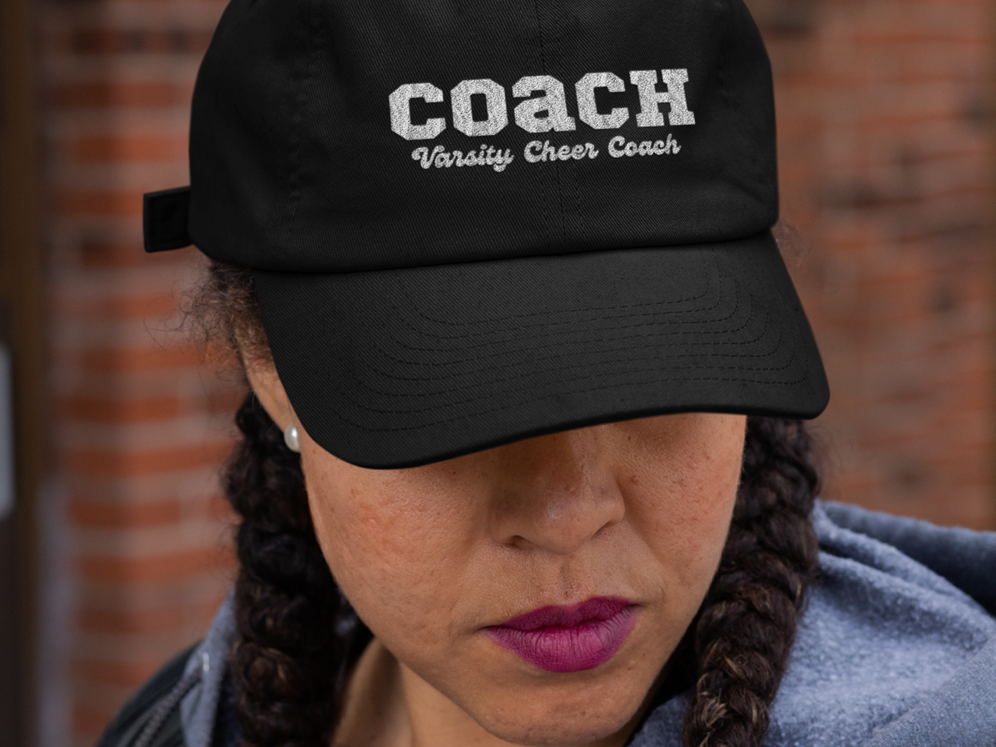 Cheerleading Coaches hat worn by a women in braids and pink lipstick in front of brick building