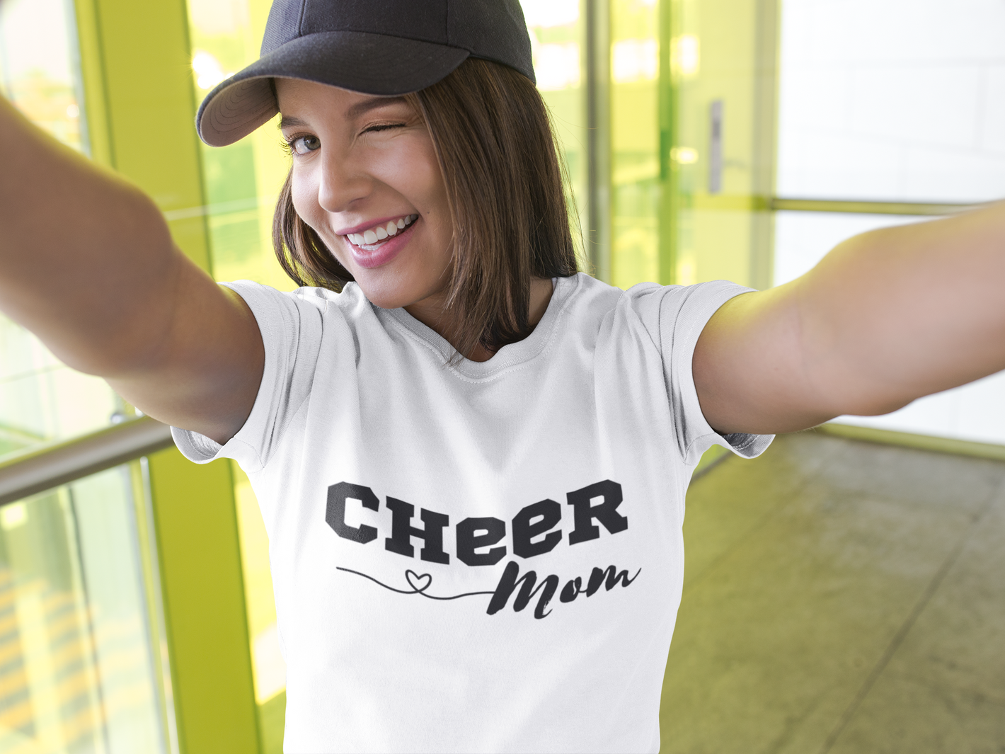 Cheerleading mom taking a selfie in front of windows wearing a t-shirt and baseball cap